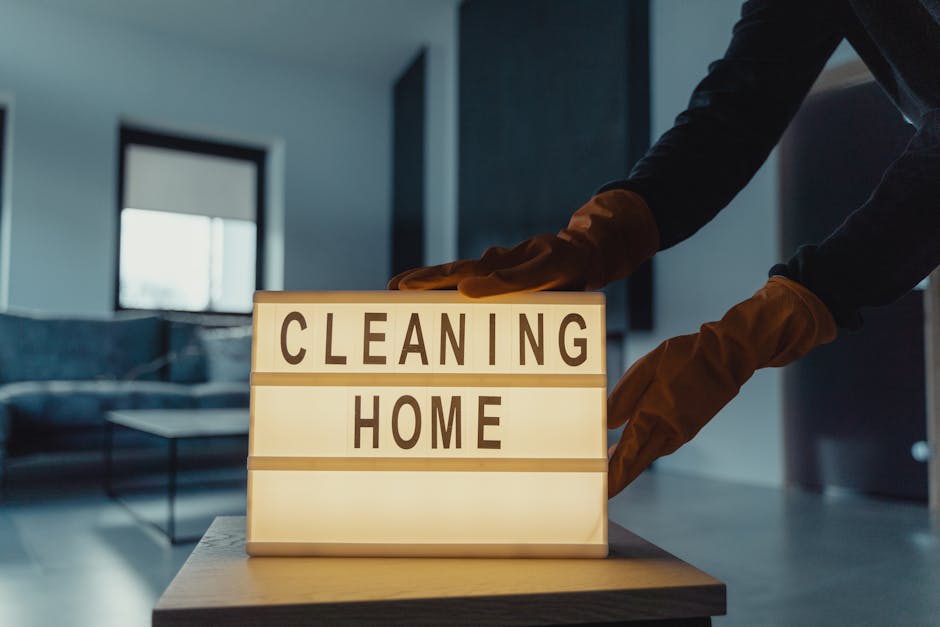 Three professional cleaners wearing grey shirts and orange overalls stand in a modern living room, each holding cleaning tools such as vacuum cleaners and mops. The room features a large window, a gre
