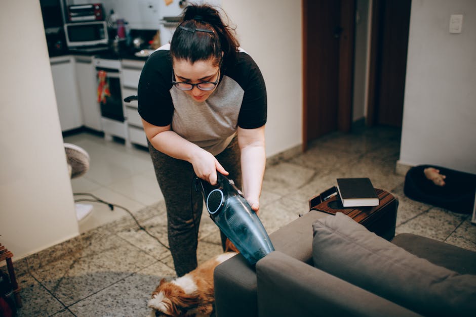 A person is vacuuming a floral patterned area rug with a yellow and black upright vacuum cleaner, executing surface cleaning within a residential living space. The person is wearing casual beige shoes