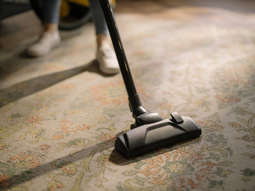 Close-up of a person using a vacuum cleaner with a black hose and floor attachment to deep clean an ornate, patterned area rug in a well-lit living room. The rug features floral motifs in muted tones 