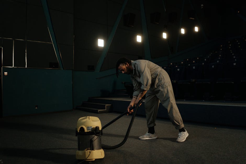 Close-up of a person using a vacuum cleaner with a black hose and floor attachment to deep clean an ornate, patterned area rug in a well-lit living room. The rug features floral motifs in muted tones 