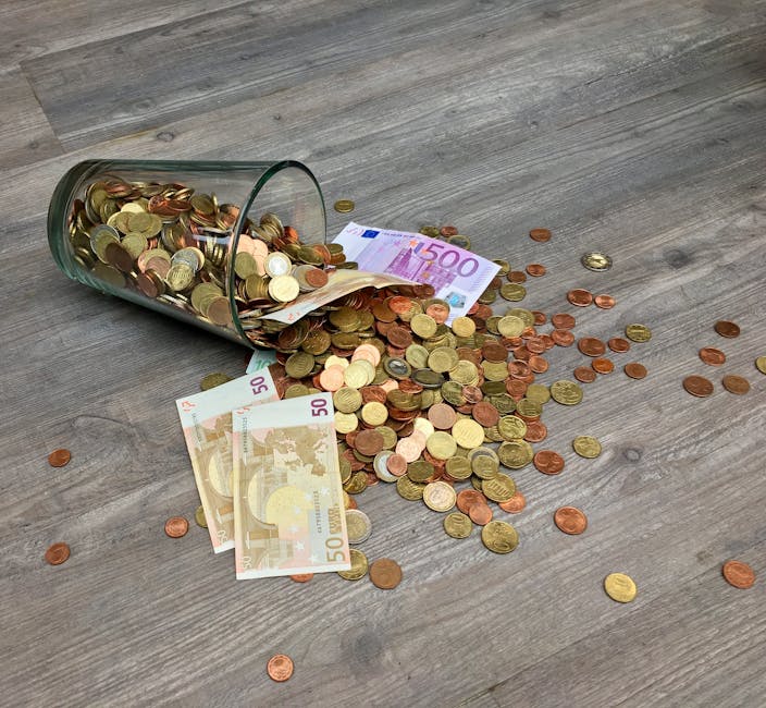 A collection of euro banknotes and assorted coins spilled onto a wooden floor. The banknotes include 50 and 500 euro bills, partially visible and crumpled among the scattered coins. The coins are of v